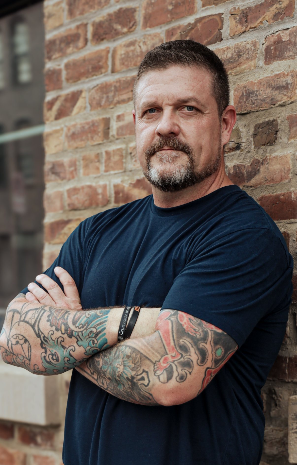Steve Baumgartner leaning on a brick wall, arms crossed, looking straight at the camera